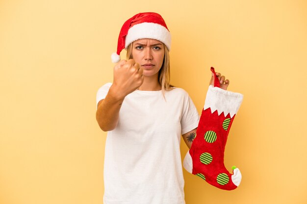 Young caucasian woman holding a elf sock isolated on yellow background showing fist to camera, aggressive facial expression.