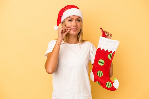 Young caucasian woman holding a elf sock isolated on yellow background with fingers on lips keeping a secret.