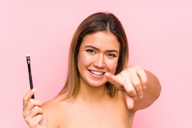 Young caucasian woman holding a eyebrush cheerful smiles pointing to front.