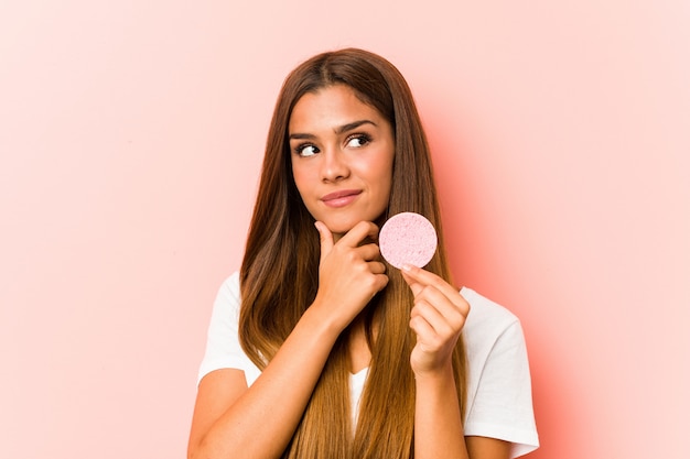 Young caucasian woman holding a facial sponge looking sideways with doubtful and skeptical expression.