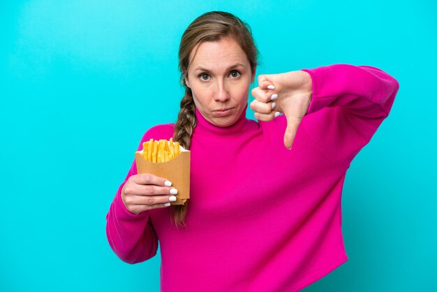 Photo young caucasian woman holding fried chips isolated on blue background showing thumb down with negative expression