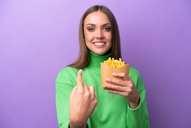 Young caucasian woman holding fried chips on purple background doing coming gesture