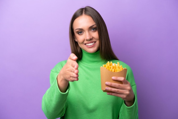 Young caucasian woman holding fried chips on purple background shaking hands for closing a good deal