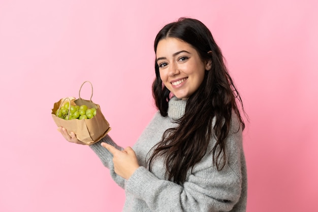 Young caucasian woman holding a grapes isolated on blue wall and pointing it