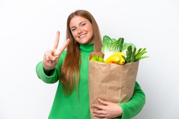 Young caucasian woman holding a grocery shopping bag isolated on white background smiling and showing victory sign