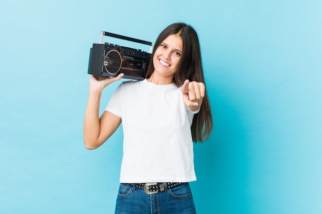 Young caucasian woman holding a guetto blaster cheerful smiles pointing to front.