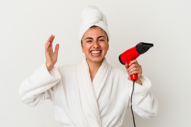 Young caucasian woman holding an hair dryer isolated on white receiving a pleasant surprise, excited and raising hands
