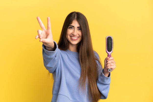 Giovane donna caucasica che tiene la spazzola per capelli isolata su fondo blu che sorride e che mostra il segno di vittoria