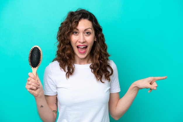 青い背景に分離されたヘアブラシを保持している若い白人女性は驚いて、人差し指を横に向けています