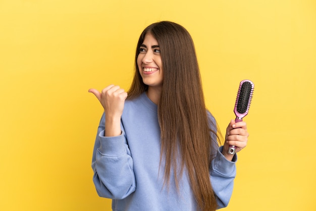 Young caucasian woman holding hairbrush isolated on blue surface pointing to the side to present a product