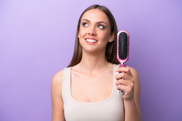 Young caucasian woman holding hairbrush isolated on purple background looking up while smiling