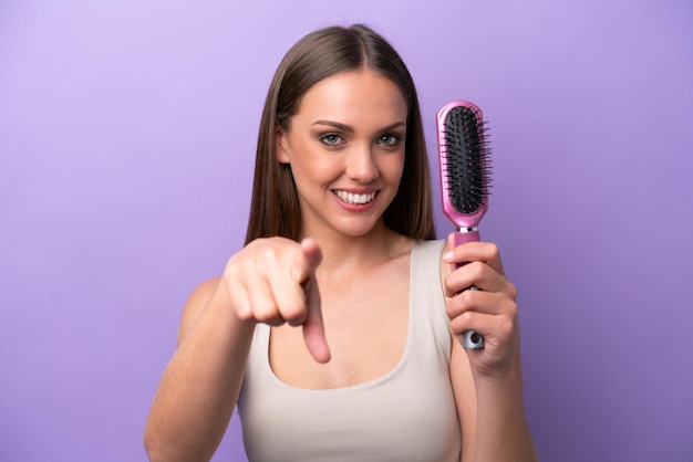 Young caucasian woman holding hairbrush isolated on purple background points finger at you with a confident expression