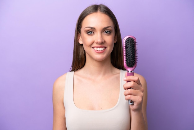 Young caucasian woman holding hairbrush isolated on purple background smiling a lot