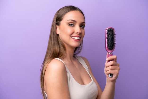 たくさんの笑みを浮かべて紫色の背景に分離されたヘアブラシを保持している若い白人女性