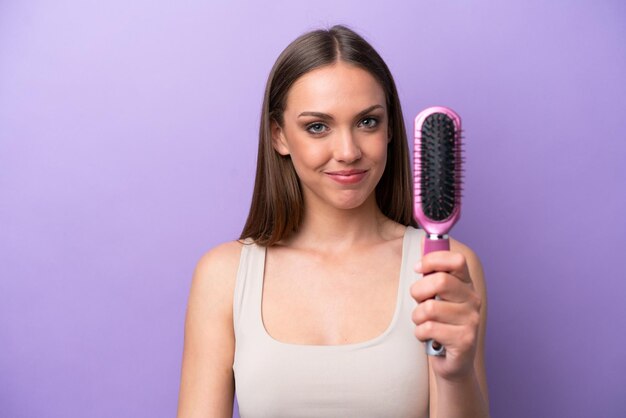 Young caucasian woman holding hairbrush isolated on purple background with happy expression