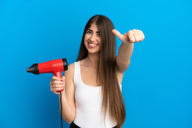 親指を立てるジェスチャーを与える青い背景で隔離のヘアドライヤーを保持している若い白人女性