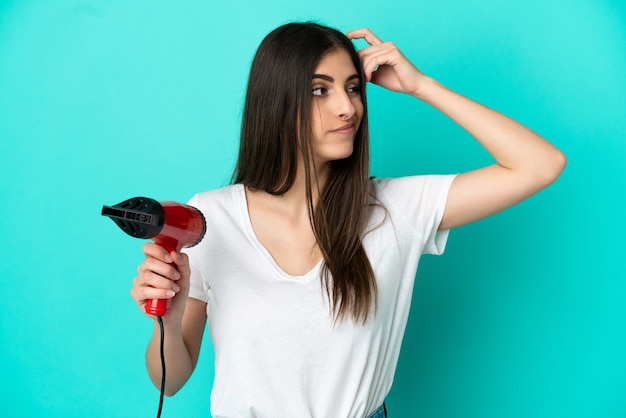 疑問と混乱した表情で青い背景に分離されたヘアドライヤーを保持している若い白人女性