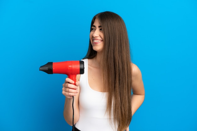 Photo young caucasian woman holding a hairdryer isolated on blue background looking side