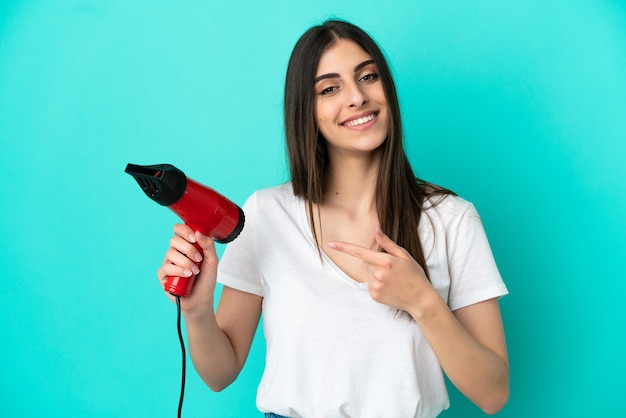 Young caucasian woman holding a hairdryer isolated on blue background and pointing it