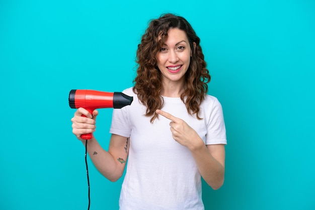 青い背景で隔離のヘアドライヤーを保持し、それを指している若い白人女性