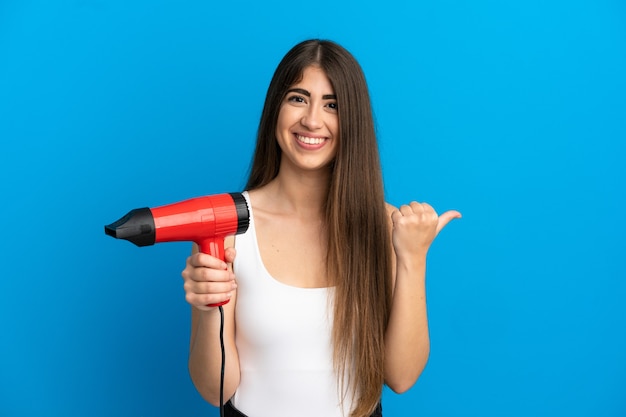 製品を提示する側を指している青い背景に分離されたヘアドライヤーを保持している若い白人女性