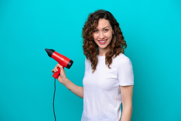 Young caucasian woman holding a hairdryer isolated on blue background smiling a lot