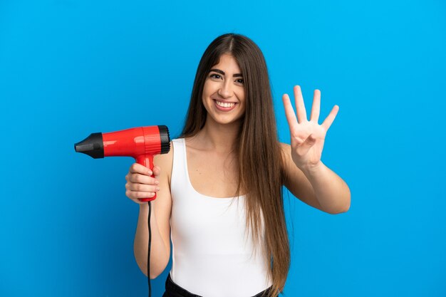 幸せな青い表面に分離されたヘアドライヤーを保持し、指で4を数える若い白人女性