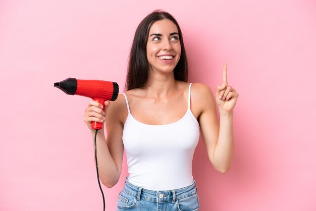 ピンクの背景に分離されたヘアドライヤーを持っている若い白人女性は、指を持ち上げながら解決策を実現しようとしています