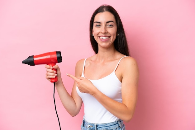 ピンクの背景に分離されたヘアドライヤーを保持し、それを指している若い白人女性