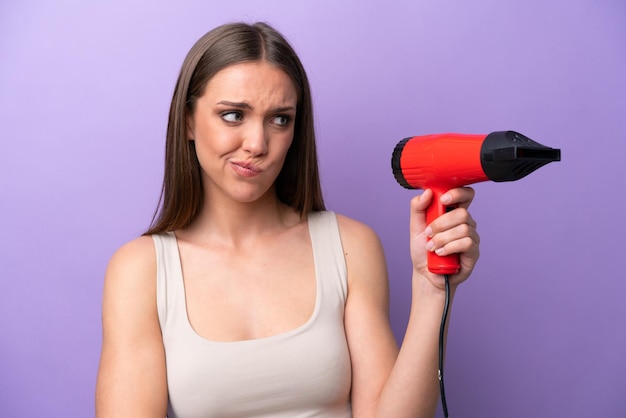 Young caucasian woman holding a hairdryer isolated on purple background with sad expression