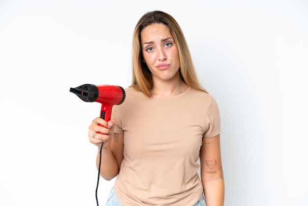 Young caucasian woman holding a hairdryer isolated on white background with sad expression