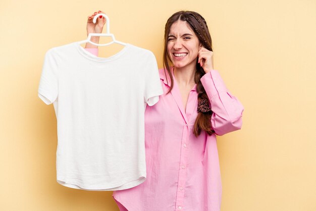 Young caucasian woman holding a hanger isolated on yellow background covering ears with hands.