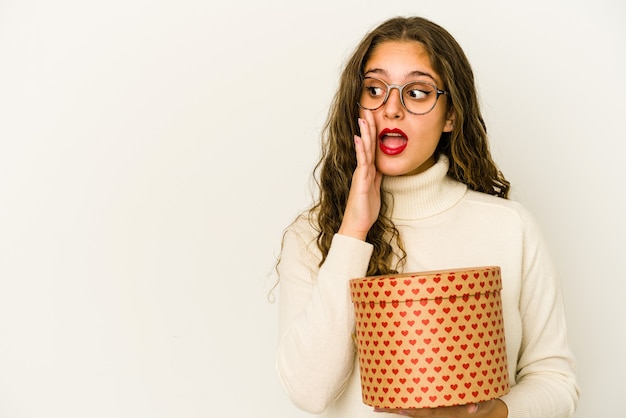 Young caucasian woman holding a heart valentines day box isolated is saying a secret hot braking news and looking aside