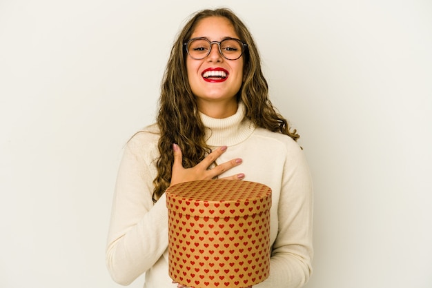 Young caucasian woman holding a heart valentines day box isolated laughs out loudly keeping hand on chest.
