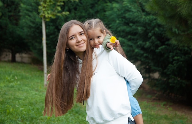 Young Caucasian woman holding her daughter on her back in the park. Upbringing concept. Mother and daughter concept.