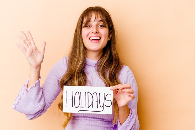 Young caucasian woman holding a Holidays placard receiving a pleasant surprise, excited and raising hands.