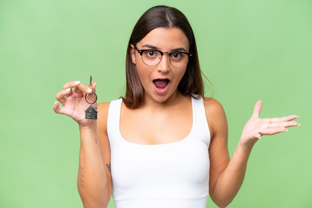 Young caucasian woman holding home keys isolated on green chroma background with shocked facial expression