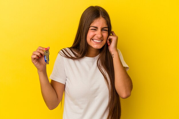 Young caucasian woman holding a home keys isolated on yellow background covering ears with hands.