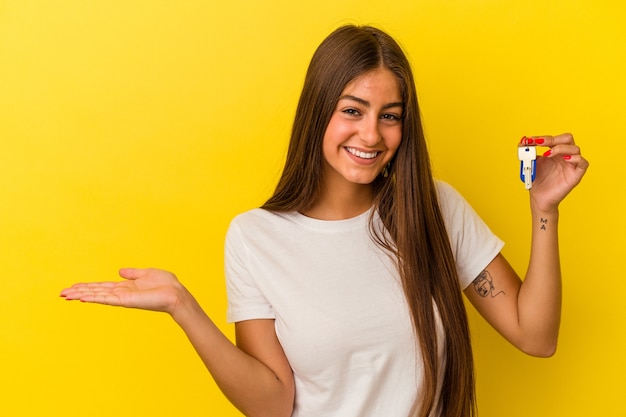 Young caucasian woman holding a home keys isolated on yellow background showing a copy space on a palm and holding another hand on waist.