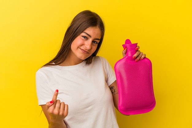 Young caucasian woman holding a hot bottle water isolated on yellow background pointing with finger at you as if inviting come closer.