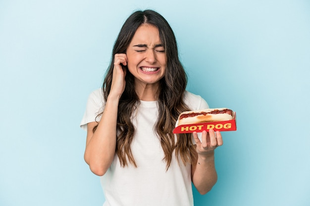 Young caucasian woman holding a hot dog isolated on blue background covering ears with hands.