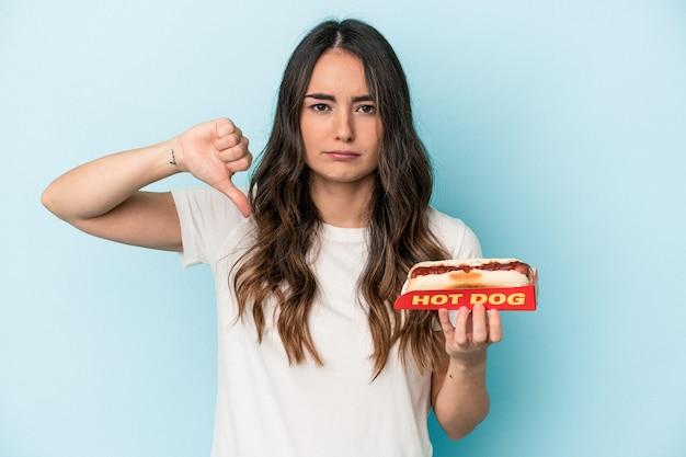 Young caucasian woman holding a hot dog isolated on blue background showing a dislike gesture, thumbs down. Disagreement concept.