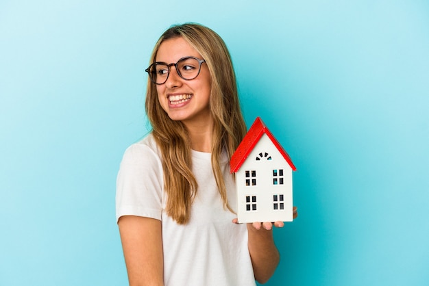 Foto la giovane donna caucasica che tiene un modello della casa isolato su priorità bassa blu guarda da parte sorridente, allegro e piacevole.