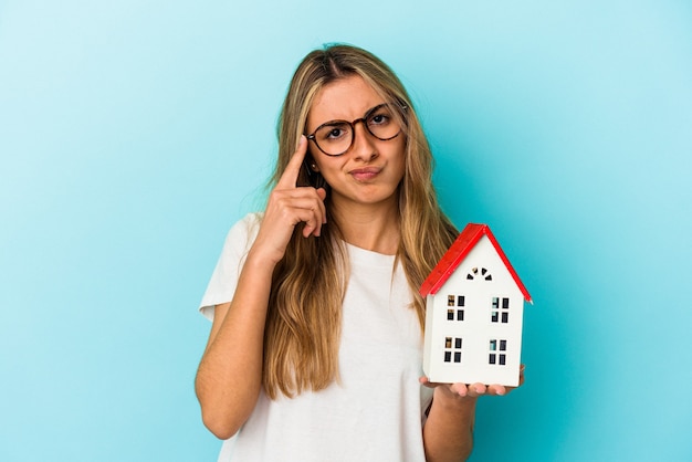 Young caucasian woman holding a house model isolated on blue background pointing temple with finger, thinking, focused on a task.