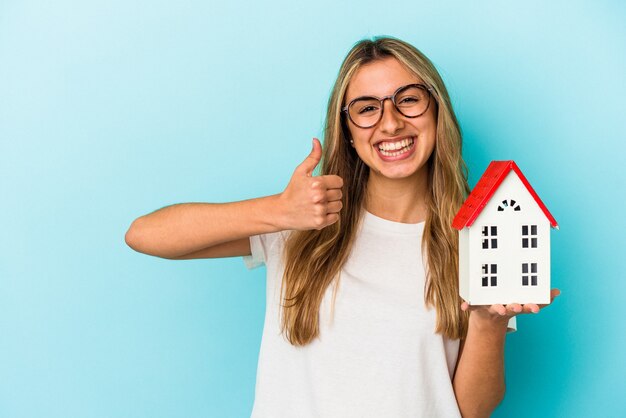 Young caucasian woman holding a house model isolated on blue background smiling and raising thumb up