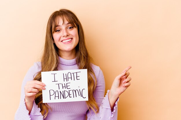 Young caucasian woman holding a I hate the pandemic placard isolated smiling and pointing aside, showing something at blank space.