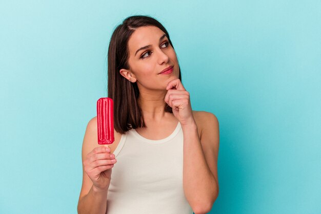 Young caucasian woman holding an ice cream isolated on blue background looking sideways with doubtful and skeptical expression.