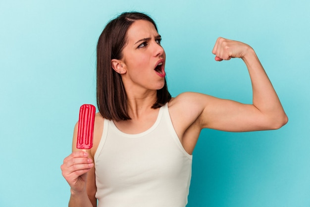 Giovane donna caucasica che tiene un gelato isolato su sfondo blu alzando il pugno dopo una vittoria, concetto vincitore.