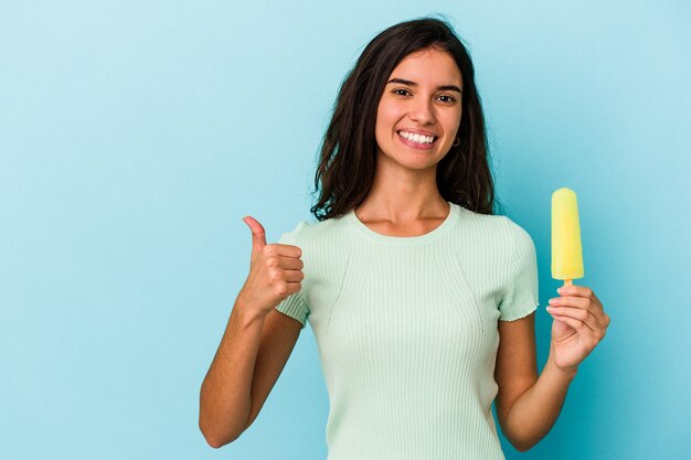 青い背景に分離されたアイスクリームを保持している若い白人女性笑顔と親指を上げる青い背景に分離されたアイスクリームを保持している若い白人女性