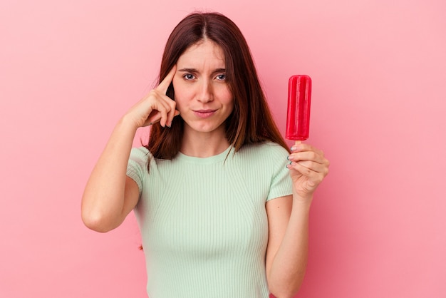 Young caucasian woman holding an ice cream isolated on blue wall pointing temple with finger, thinking, focused on a task.